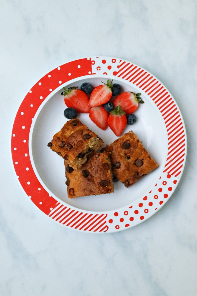 3 squares of Healthier Chocolate Chip Cake Bar on a red and white plate with sliced strawberries and blueberries.