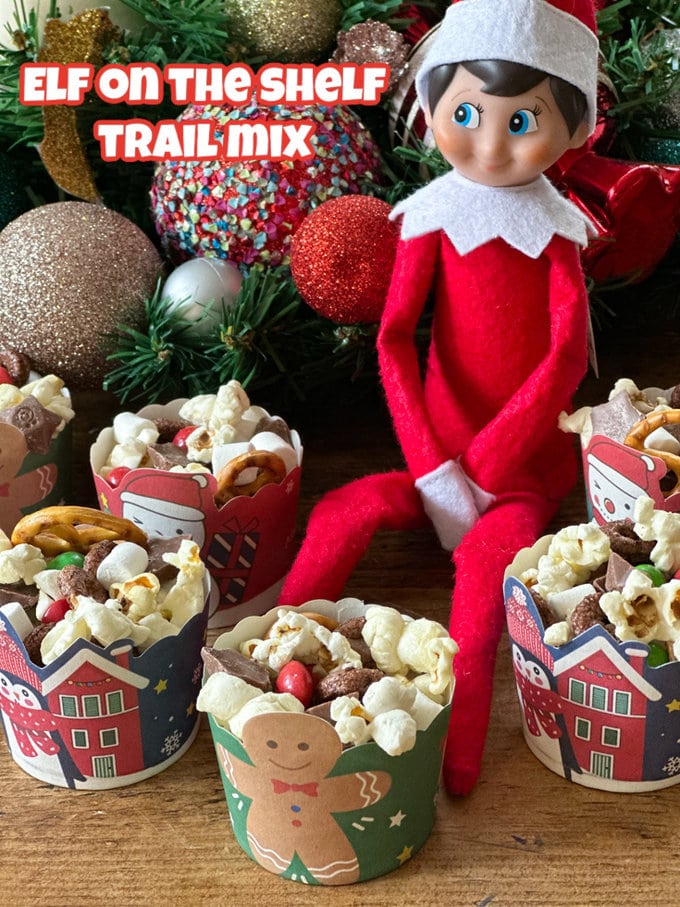 Small Christmas themed muffin cups filled with Christmas Trail Mix with an Elf On The Shelf sitting amongst them with Christmas decorations in the background.