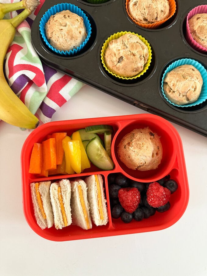 3 Ingredient Banana Muffins in a muffin tray with a lunchbox in the foreground containing a muffins, a sandwich and some fresh chopped fruit and veggies.
